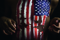 In this extreme close-up of body paint on a torso, a Black person’s chest is painted with a peeling image of the American flag, aligned vertically. The field of blue on the flag, which appears on the upper left side of the chest, has white lines to represent stars. The figure whose body is painted appears to be in the posture of a runner, elbows bent and the hands curved in tension.
