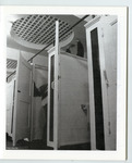 Fig. 124. Black-and-white photograph of the inside of a public restroom, with white stalls and two individuals inside. We see only the arms of one, placed against the ceiling, and part of the back of another, suggesting a sexual encounter between them.
