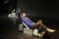 A female dancer is lounging on a series of electronic devices such as computer screens and printers positioned to resemble furniture. In the back an out-­of-­focus male dancer does the same.