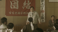 A man stands in front of an assembled class, with a large red banner and vertical scroll of calligraphic text behind him