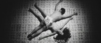 A naked man and woman lie across each other diagonally in a spotlight on a floor covered in paper saturated with calligraphy, in black and white cinematography.