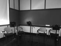 Black and white photo, four chairs are positioned in front of iPads mounted on stands. There are two speakers and some cables on the floor in the back.