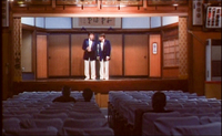 Two men perform on a stage to a mostly empty crowd, surrounded by various banners with black calligraphy.