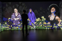 Photo: A large digital installation. There is one wall with a video being projected on it that gives the visual effect of layers. The background layer is a contrast of black with large white petals from a cotton plant. In the foreground are purple-and-yellow poppies and yellow roses. In the middle, there are three Black women of different ages, each wearing a different garment to represent the time period she represents. In front of the video projection is a viewer looking at the artwork.