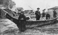 A black and white photograph of five indigenous men and women standing in or around a long dugout canoe. Written text in white on the bottom of the photograph reads, "Indian Family near Chimacum Creek, Wash."