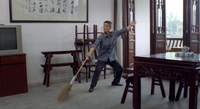 A man extends a broom in a restaurant as if he's dancing. The wall behind him has a large, horizontal work of framed calligraphy.