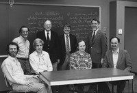 Figure 2. Eight white male scientists sit and stand in two rows in front of a chalkboard covered in math problems.