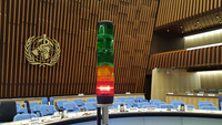 Picture of a cylindric traffic light. Three green lights are at the top, followed first by an orange light and then by a red light. The red light is currently on. In the background, the emblem of the World Health Organization and seats for meeting participants can be seen.