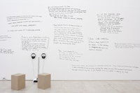 Photograph of an art gallery wall inscribed from floor to ceiling with handwritten testimony (mostly indecipherable). A listening station, with two headphones and wooden seats, sits at the bottom left of the image.