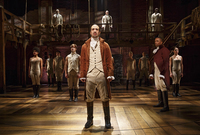 Alexander Hamilton surrounded by ensemble members wearing black boots and beige costumes that include waistcoats in velvet textures and corseted vests with no sleeves.