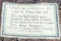 A large rectangular tile plaque with a painted border, set into a stone wall. The painted words on the plaque read as follows: “Aquí se estableció en 1700 el Hospital del Divino Salvador para mujeres dementes, fundado en el siglo XVII por el carpintero José Sayago. —Dirección de monumentos coloniales de la república.” [In 1700, the Hospital of the Divine Savior for insane women, founded in the seventeenth century by the carpenter José Sayago, was established here. —Administration of colonial monuments of the republic].