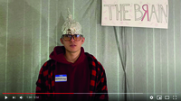 Color image of one frame from video showing student with aluminum foil cap and “Dopamine” name tag, standing in front of plain gray background. Sign behind student says, “The Brain” with letter “r” written backward.