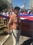 A Black person in their drag persona. They wear a painted-on moustache and a button-down, short-sleeved shirt and tie, and they hold up part of the Cuban flag for a march.