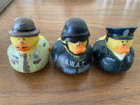 Photograph of three rubber ducks in various police costumes. On the left is a rubber duck in a green and brown sheriff’s costume and hat, in the center a rubber duck in a SWAT team uniform and helmet, and on the right a rubber duck in a navy-­blue police costume and cap.