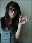 A white woman with long dark hair and thick glasses smirks at the camera and shows off the word “NERD” written on the palm of her hand.