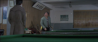 The male protagonist stands in the left of the frame, in front of one pool table in his trench coat in the foreground. Two banners are posted on the wall to his left, one with white calligraphy on a blue matte and the other with black calligraphy on a white matte. The protagonist looks at another man who looks back at him from a second pool table in the midground, holding a pool cue. A sign is posted on the wall above the second man.