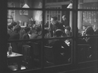 A view of a classroom of young men sitting around a table has white calligraphy written on a chalkboard and black calligraphy on various banners on the wall, in black and white cinematography.
