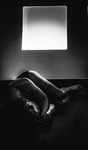 Black-and-white photograph of a female figure lying on the floor in fetal position in the shadow, below a reflection of light on the wall.