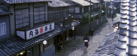 Image of a street lined with shops and a single man in kimono walking in the middle of the street. The calligraphic sign on the left-hand store indicates iit is a rice shop.