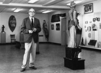 Black and white photo showing a person standing in a room containing sculptures and other artistic works.