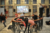 A metal banister structure is in the middle of a large room. Dancers are climbing over and hanging from the structure. A screen attached to the back wall shows people holding hands at a social gathering.
