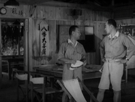 Two men talk to each other, inside a wooden building. Calligraphy is visible on banners and signs inside.