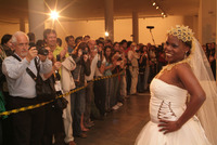 A Black trans person in a wedding dress with a veil cap made of condoms poses, smiling, for a crowd of photographers.