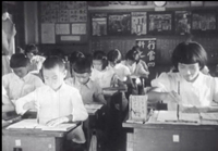 A classroom of pupils sit in front of a banner with white calligraphy, in black and white cinematography.