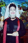 Mercado is grinning, has a white cloth on head with a black braid, and is holding a maraca in one hand and a folded fan in the other.