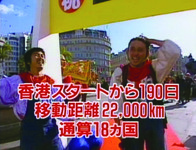 Color still. Two men (duo Saruganseki) stand before finish line banner. Men are wearing blue overalls and white shirts while gesturing relief. On screen, Japanese text reads: “190 days from start in Hong Kong; Travel distance 22,000 km; 18 countries total.”