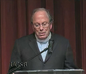Image of a man speaking at a podium in front of a red curtain. A watermark with the letters UCSB is in the lower left corner.