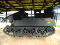 A black metal tank stands on a concrete floor under a corrugated iron roof. Each continuous metal track passes around six wheels, and a Biafran flag adorns the back of the tank. Several other large, industrial war machines are visible in the background.