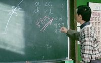 An instructor draws calligraphy and English letters on a chalkboard. A poster with a series of boxes with green calligraphy is posted on the wall behind him.