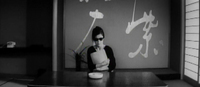 A woman sits at a low table, holding papers, in front of white calligraphy on the wall.