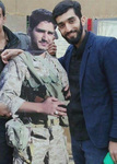 Mohsen Hojaji, an Iranian military officer, stands alongside a life-­sized cutout of a martyred soldier wearing army fatigues. Hojaji is wearing dark blue trousers, a dark blue jacket, and a black shirt, and stands a little taller than the martyr.