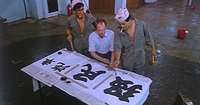 In a high angle shot protagonist Mark paints over black calligraphy of a store name on white paper, as two others observe him.