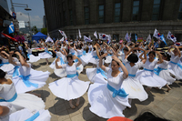 Figure 15.1. A group of Korean women in white dresses and blue waist sash raise their arms above heads as they twirl. Behind them, protesters wave the South Korean national flags.