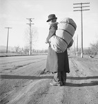 Fig. 22. A man in a frayed overcoat and dusty shoes stands on the dirt shoulder beside an empty road. He carries a large sack slung over his shoulder.