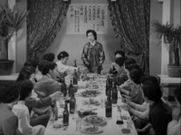 A group of women sit at a dining table in front of a poster on the wall with black calligraphy, in black and white cinematography.
