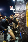 Artist Xandra Ibarra, wearing a see-through, long-sleeve top, a strap-on harness, and knee-high fishnet socks, faces and leans into a brick wall, while a person standing behind her prepares to put paper money somewhere on her.