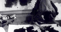A woman bows painting large black calligraphy on paper, in black and white cinematography.
