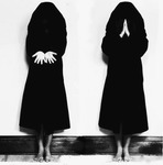 Black-and-white photograph of two identical figures dressed in a dark cloak side by side.