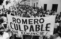 Photo of a banner that names the governor and says “Romero Guilty, Resign”; behind, a multitude fills the street leading to the governor’s official residence.