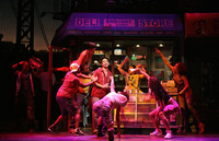 Usnavi surrounded by eight dancers as he sings “96,000” in front of the bodega. One dancer has a boom box on his shoulder.