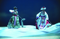A color photo of a scene on stage. Two female figures are controlling two snake puppets, holding a stick in each hand. The figure on the right wears a headpiece and is in a white robe and is controlling a white snake puppet, the figure on the left wears a green robe and is controlling a green snake puppet. The stage is covered with a giant piece of blue silk that represents the raging waves.