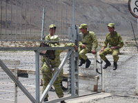 A small group of border guards sit along a metal fence that separates the Tajik side of the border from the cross-border bazaar. They are laughing and smiling. There are people in the background and an entrance in the fence for people to access the bazaar. Also in the background are a bridge that crosses the river and the low-lying mountains across the river in Afghanistan.