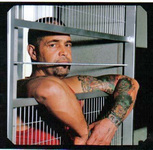 A bearded man with heavily tattooed arms leans against the insubstantial bars of his mock prison cell, looking directly toward the camera.