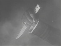 In black and white cinematography, a close-up of a graphic shows a man's hand holding a knife at the beginning of the film. There is calligraphy on the knife, reading katakana カオ at the end of the shaft. The hand is wearing a ring with the Japanese Imperial Flag.