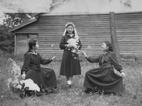 In this photograph from the early 1950s, a young Japanese woman stands outside posing in pretend bridal attire, enacting an American-­style wedding, while her two friends present her with flowers.