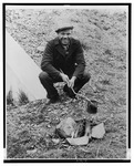 Fig. 30. A young man in dark, casual clothing and a sports cap holds a tin can on a stick over a fire. He is squatting outside, next to what looks like a tent.